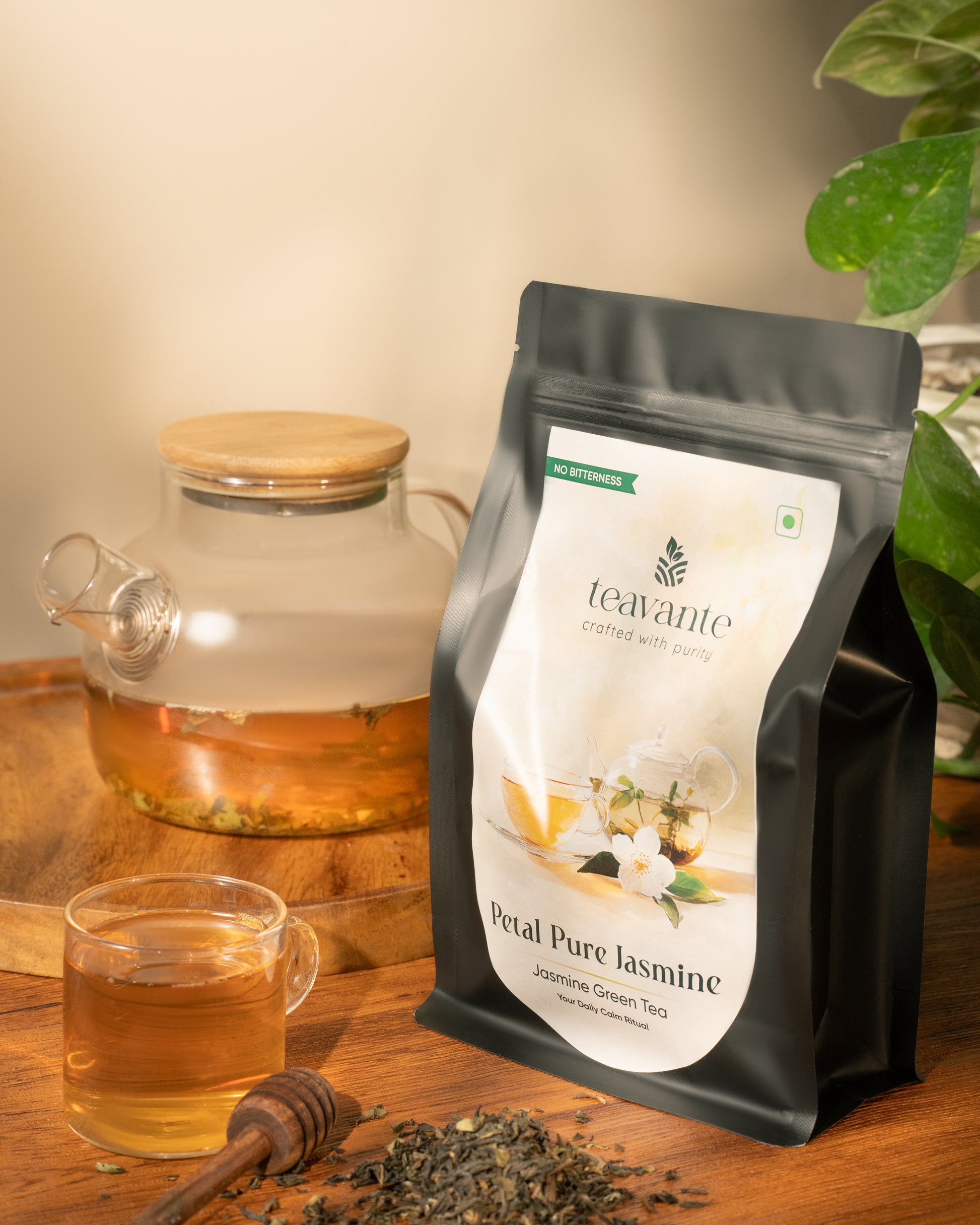 A stand-up pouch of Teavante Petal Pure Jasmine organic green tea stands on a wooden table beside a glass teapot, a glass cup of amber tea, loose tea leaves, a honey dipper, and a plant leaf. The pouch label shows the Teavante brand and product name.